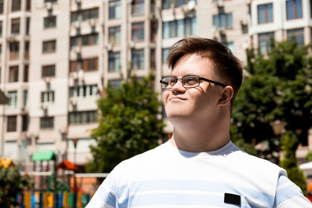 A smiling young man with Down syndrome in glasses poses against the background of the city