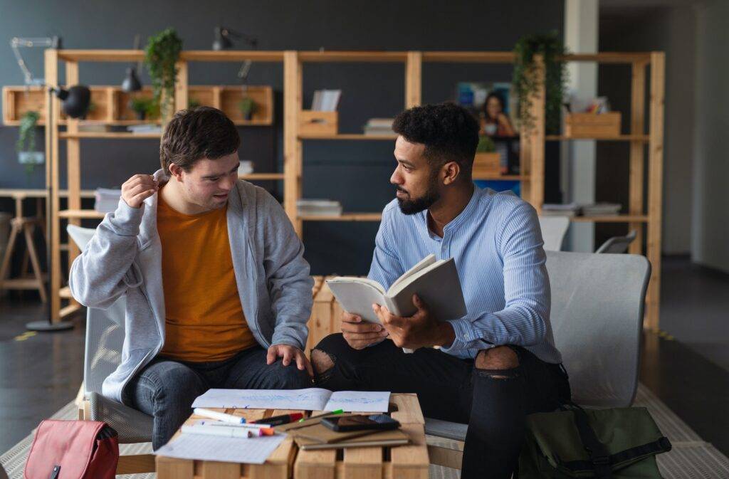 Young happy man with Down syndrome and his tutor studying indoors at school